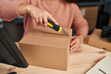 Young hispanic woman ecommerce business worker unpacking cardboard box using cutter at office