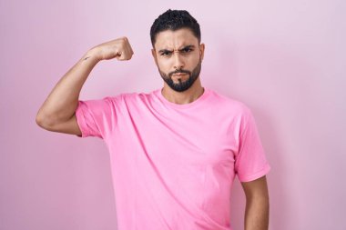 Hispanic young man standing over pink background strong person showing arm muscle, confident and proud of power 