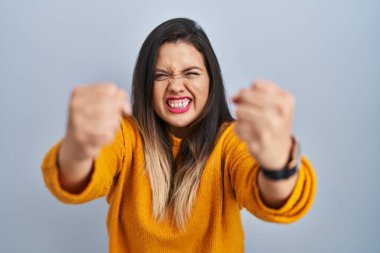 Young hispanic woman standing over isolated background angry and mad raising fists frustrated and furious while shouting with anger. rage and aggressive concept. 