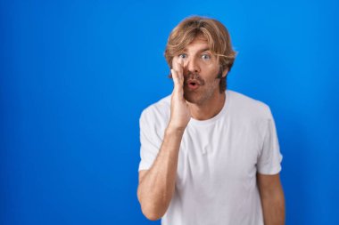 Middle age man standing over blue background hand on mouth telling secret rumor, whispering malicious talk conversation 