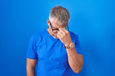 Hispanic man with grey hair standing over blue background tired rubbing nose and eyes feeling fatigue and headache. stress and frustration concept. 