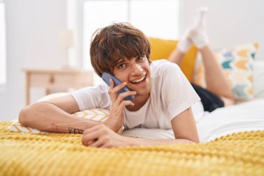 Genç sarışın adam yatakta uzanmış akıllı telefondan konuşuyor.