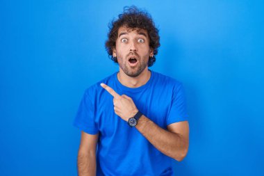 Hispanic young man standing over blue background surprised pointing with finger to the side, open mouth amazed expression. 