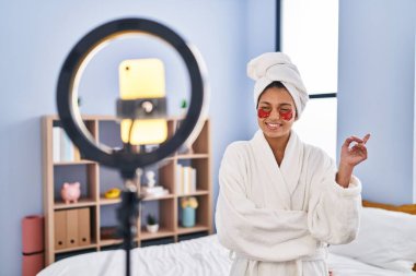 Koyu renk saçlı İspanyol kadın akıllı telefonuyla güzellik dersi kaydediyor. Gülümsüyor, mutlu bir şekilde elini ve parmağını yana doğru gösteriyor. 