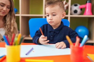 Teacher and toddler sitting on table drawing on paper at kindergarten