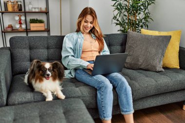 Genç beyaz kadın dizüstü bilgisayar kullanıyor. Kanepede köpeğiyle oturuyor.