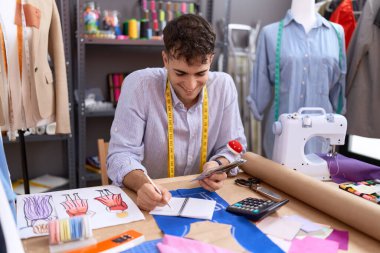 Young hispanic man tailor holding dollars writing on notebook at atelier