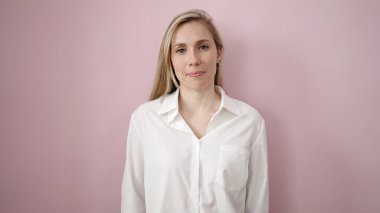 Soyutlanmış pembe arka planda ciddi bir ifadeyle duran genç sarışın kadın.