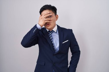 Young asian man wearing business suit and tie peeking in shock covering face and eyes with hand, looking through fingers with embarrassed expression. 