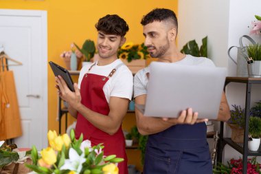 Two hispanic men florists using laptop and touchpad working at flower shop