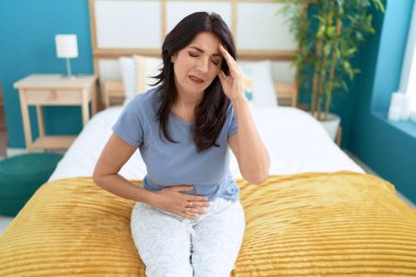 Middle age hispanic woman suffering for head and stomach ache sitting on bed at bedroom