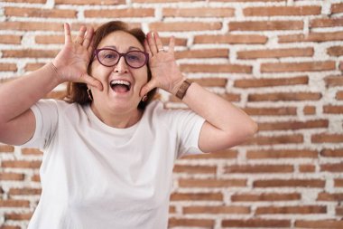 Senior woman with glasses standing over bricks wall smiling cheerful playing peek a boo with hands showing face. surprised and exited 
