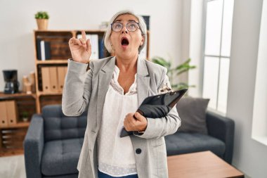 Middle age woman with grey hair at consultation office amazed and surprised looking up and pointing with fingers and raised arms. 