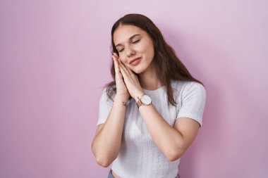 Young hispanic girl standing over pink background sleeping tired dreaming and posing with hands together while smiling with closed eyes. 