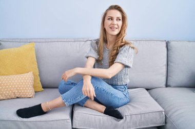 Sarışın genç bir kadın evdeki koltukta kendinden emin bir şekilde oturarak gülümsüyor.