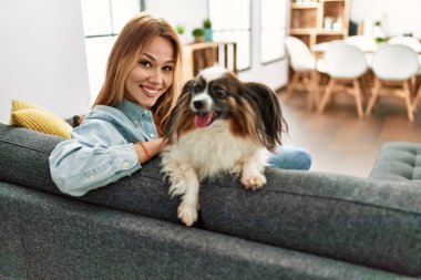 Beyaz bir kadın, evinde köpekle kanepede otururken kendinden emin bir şekilde gülümsüyor.