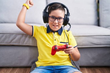 Young hispanic kid playing video game holding controller wearing headphones angry and mad raising fist frustrated and furious while shouting with anger. rage and aggressive concept. 