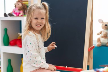 Adorable blonde girl preschool student drawing on blackboard at kindergarten