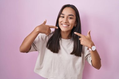 Pembe arka planda duran, neşeli gülümseyen ve parmaklarıyla ve ağzıyla işaret eden genç İspanyol kadın. diş sağlığı kavramı. 