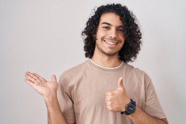 Hispanic man with curly hair standing over white background showing palm hand and doing ok gesture with thumbs up, smiling happy and cheerful 