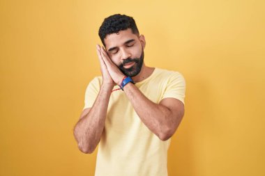 Hispanic man with beard standing over yellow background sleeping tired dreaming and posing with hands together while smiling with closed eyes. 