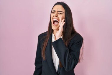 Young brunette woman wearing business style over pink background shouting and screaming loud to side with hand on mouth. communication concept. 