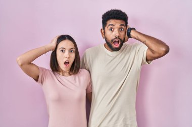 Young hispanic couple together over pink background crazy and scared with hands on head, afraid and surprised of shock with open mouth 