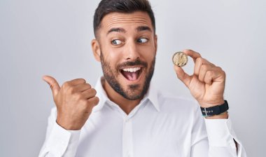 Handsome hispanic man holding litecoin cryptocurrency coin pointing thumb up to the side smiling happy with open mouth 