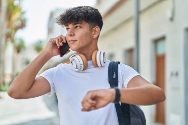 Young hispanic teenager student talking on smartphone looking watch at street