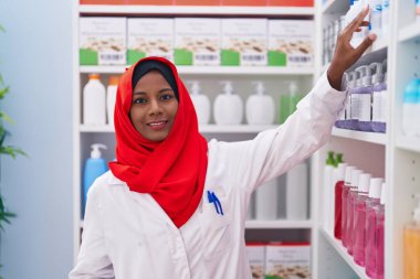 Young beautiful woman pharmacist smiling confident holding medication on shelving at pharmacy