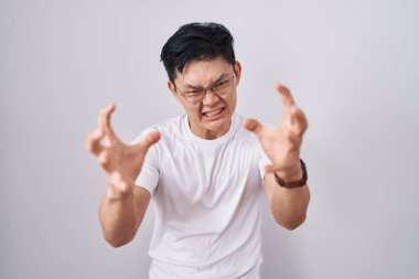 Young asian man standing over white background shouting frustrated with rage, hands trying to strangle, yelling mad 