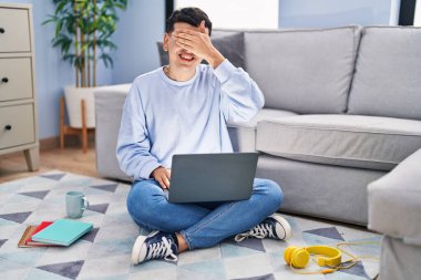 Non binary person studying using computer laptop sitting on the floor smiling and laughing with hand on face covering eyes for surprise. blind concept. 