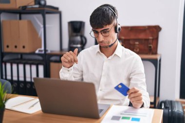 Young hispanic man working using computer laptop holding credit card pointing down looking sad and upset, indicating direction with fingers, unhappy and depressed. 