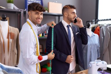 Two hispanic men tailor measuring sleeve jacket while client talk on smartphone at atelier