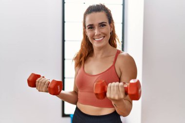 Genç kızıl saçlı kadın gülümsüyor spor merkezinde kendine güvenen dambıllarla antrenman yapıyor.