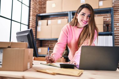 Young beautiful hispanic woman ecommerce business worker using laptop and calculator writing on notebook at office