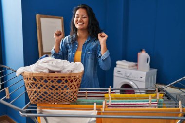 Young asian woman hanging clothes at clothesline excited for success with arms raised and eyes closed celebrating victory smiling. winner concept. 