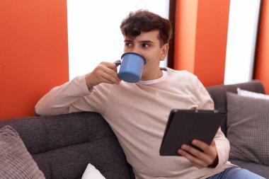 Non binary man using touchpad drinking coffee sitting on sofa at home