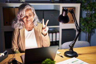 Middle age woman with grey hair working using computer laptop late at night showing and pointing up with fingers number two while smiling confident and happy. 