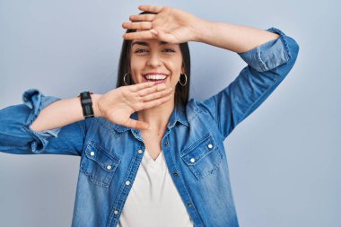 Hispanic woman standing over blue background smiling cheerful playing peek a boo with hands showing face. surprised and exited 