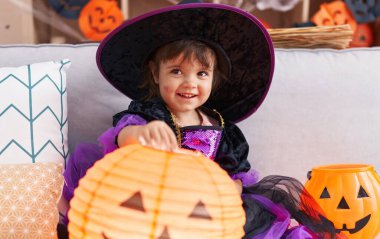 Adorable hispanic girl wearing halloween costume holding pumpkin basket lamp at home