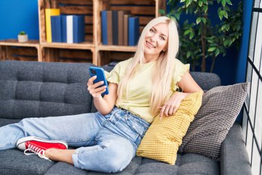 Evdeki kanepede oturan akıllı telefon kullanan genç sarışın kadın.