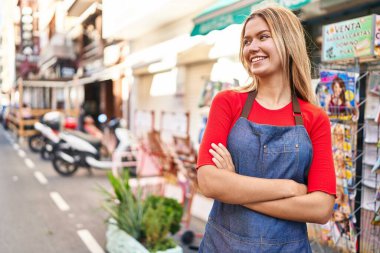 Young hispanic woman shop assistant standing with arms crossed gesture at street