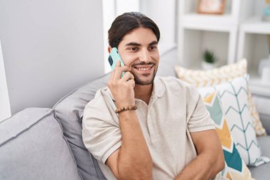Genç İspanyol adam akıllı telefonda konuşuyor. Evdeki kanepede oturuyor.