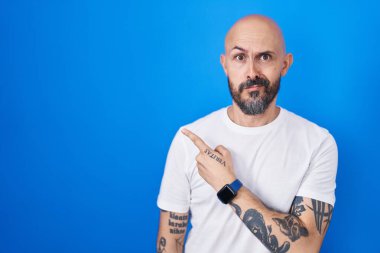 Hispanic man with tattoos standing over blue background pointing aside worried and nervous with forefinger, concerned and surprised expression 