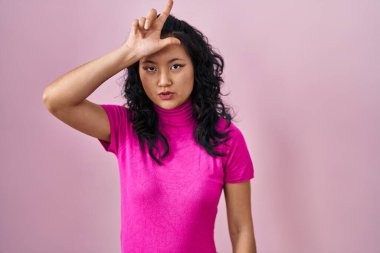 Young asian woman standing over pink background making fun of people with fingers on forehead doing loser gesture mocking and insulting. 