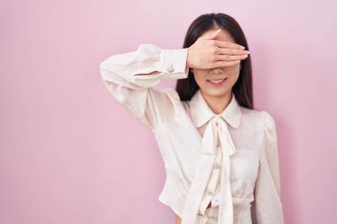 Young chinese woman standing over pink background smiling and laughing with hand on face covering eyes for surprise. blind concept. 