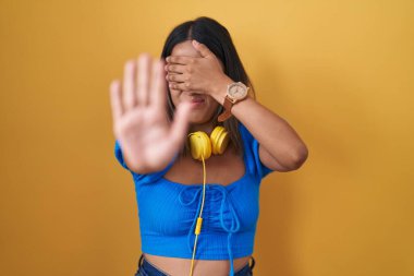 Hispanic young woman standing over yellow background covering eyes with hands and doing stop gesture with sad and fear expression. embarrassed and negative concept. 