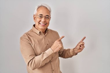Hispanic senior man wearing glasses smiling and looking at the camera pointing with two hands and fingers to the side. 