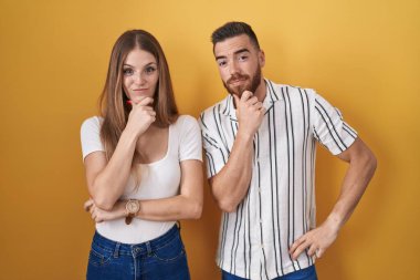 Young couple standing over yellow background looking confident at the camera smiling with crossed arms and hand raised on chin. thinking positive. 